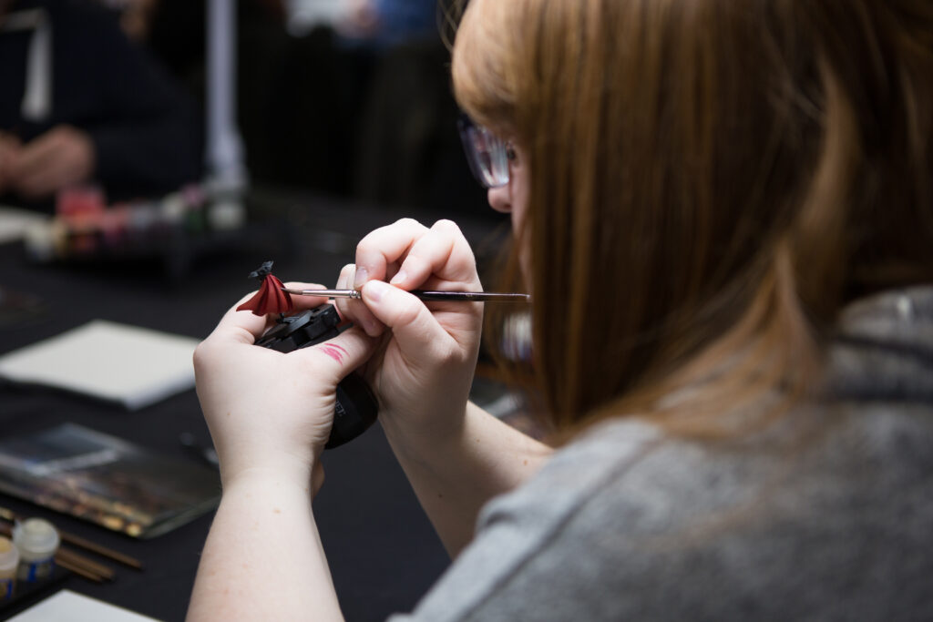 A young woman with glasses is shown carefully painting her Warhammer 40,000 miniatures. She is seated at a desk, surrounded by brushes, paints, and small figurines. The focus of the image is on her concentration and skill as she brings life to her army with each stroke of her brush. 